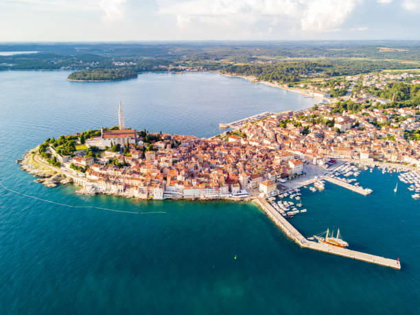 Croatian town of Rovinj on a shore of blue azure turquoise Adriatic Sea, lagoons of Istrian peninsula, Croatia. High bell tower, red tiled roofs of historical buildings, sailboat, piers. Aerial view.