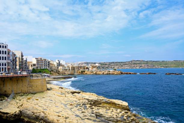 View along St Pauls Bay waterfront and rocky coastline with apartments to the left and people going enjoying the setting, Bugibba, Malta, Europe.